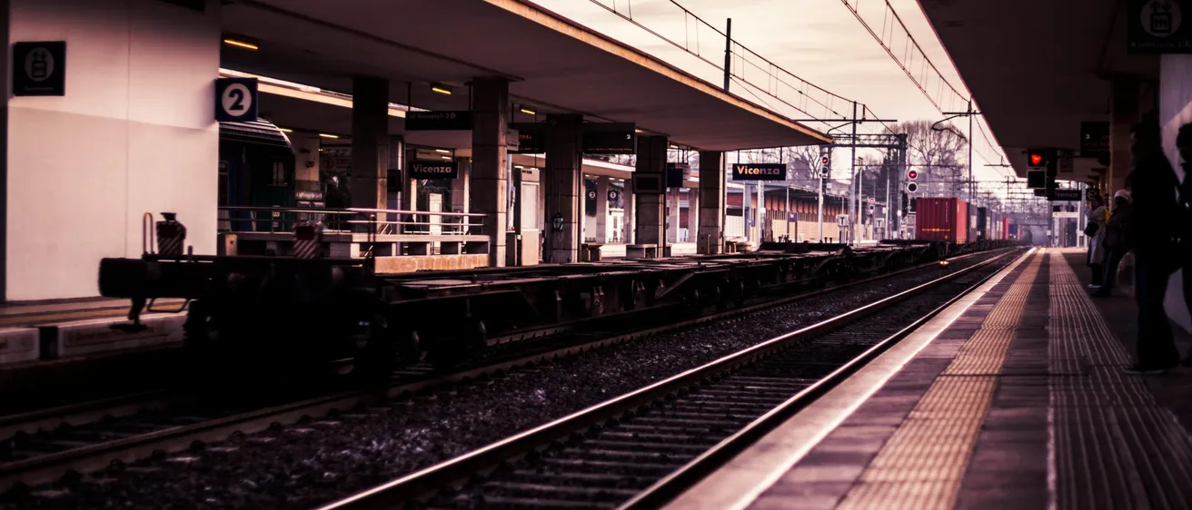 Ein Bahnhof im Freien mit Bahnsteig, Oberleitungen und einem Zug auf der Strecke.