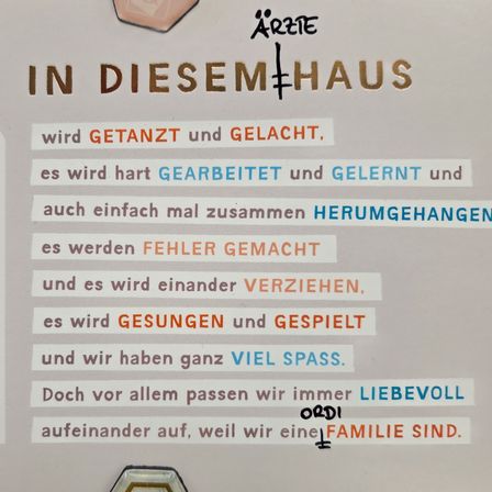 Ein Plakat mit verschiedenen Phrasen auf Deutsch. Oben steht 'IN DIESEM+HAUS'. Darunter sind Phrasen wie 'wird GETANZT und GELACH,' 'es wird hart GEARBEITET und GLERNT' und 'es werden FEHLER GEMACHT.' Das Plakat betont Familienliebe und Einheit.