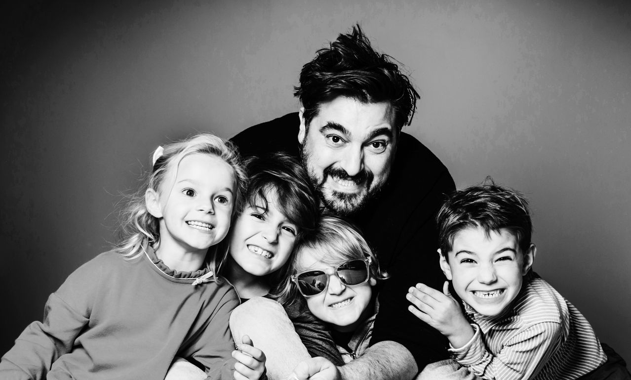 Ein schwarz-weißes Familienporträt mit einem bärtigen Vater und vier lächelnden Kindern, die eng bei ihm stehen.