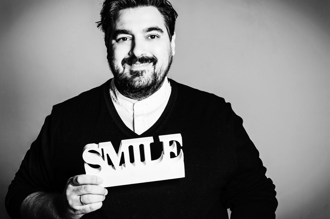 Ein Mann mit Bart lächelt und hält ein weißes Schild mit der Aufschrift 'SMILE' vor seiner Brust. Er trägt einen schwarzen Pullover.