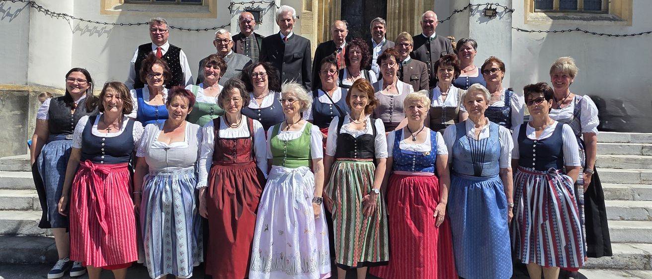 Eine Gruppe von Menschen in traditioneller Kleidung, sowohl Männer als auch Frauen, steht vor einem historischen Gebäude mit einem Bogeneingang. Sie alle lächeln für ein Gruppenfoto.