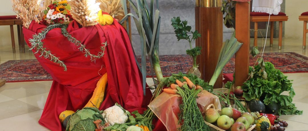 Ein Altar mit einem hölzernen Kreuz und einem Weizenkranz liegt vor einem geschmückten Altar, umgeben von einer Vielfalt an Gemüse und Früchten auf einem roten Tuch.