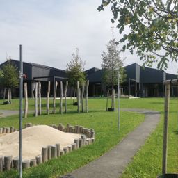 Ein Rasenfeld mit einem Gehweg führt zu einer Reihe schwarzer Gebäude mit Solarpaneelen. Das Gelände ist von jungen Bäumen und einem Sandkasten umgeben.