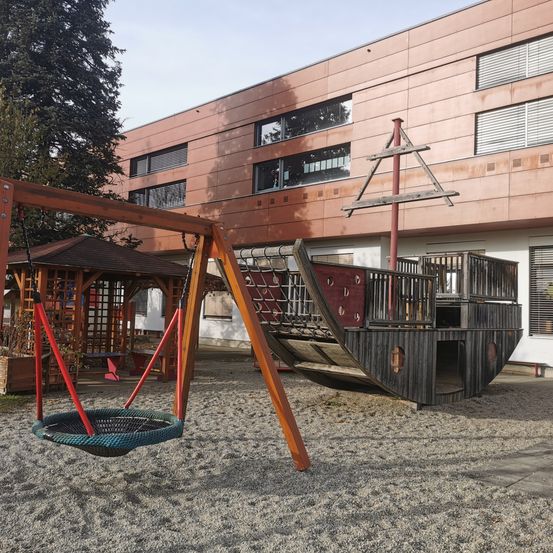 Ein Spielplatz mit einem Piratenschiff und einer Schaukel, vor einem modernen Gebäude mit mehreren Fenstern gelegen.