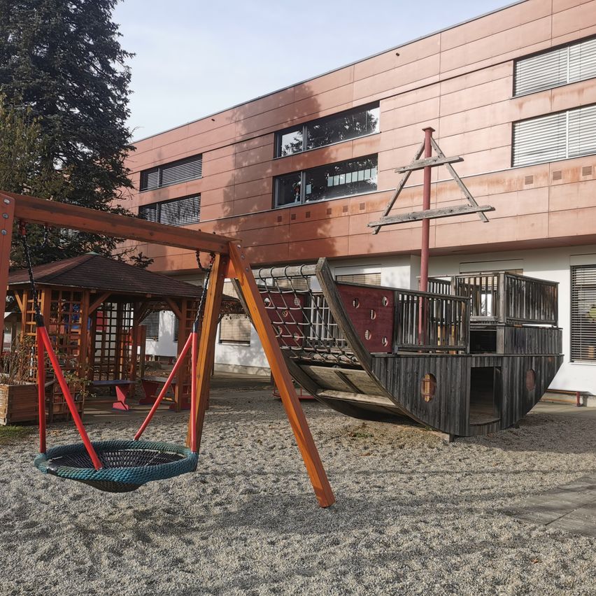 Ein Spielplatz mit einem Piratenschiff und einer Schaukel vor einem modernen Gebäude mit vielen Fenstern.