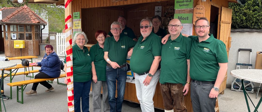 Eine Gruppe von Menschen in grünen Hemden und Hosen posiert für ein Foto vor einem hölzernen Stand mit Schildern und einer Menütafel.