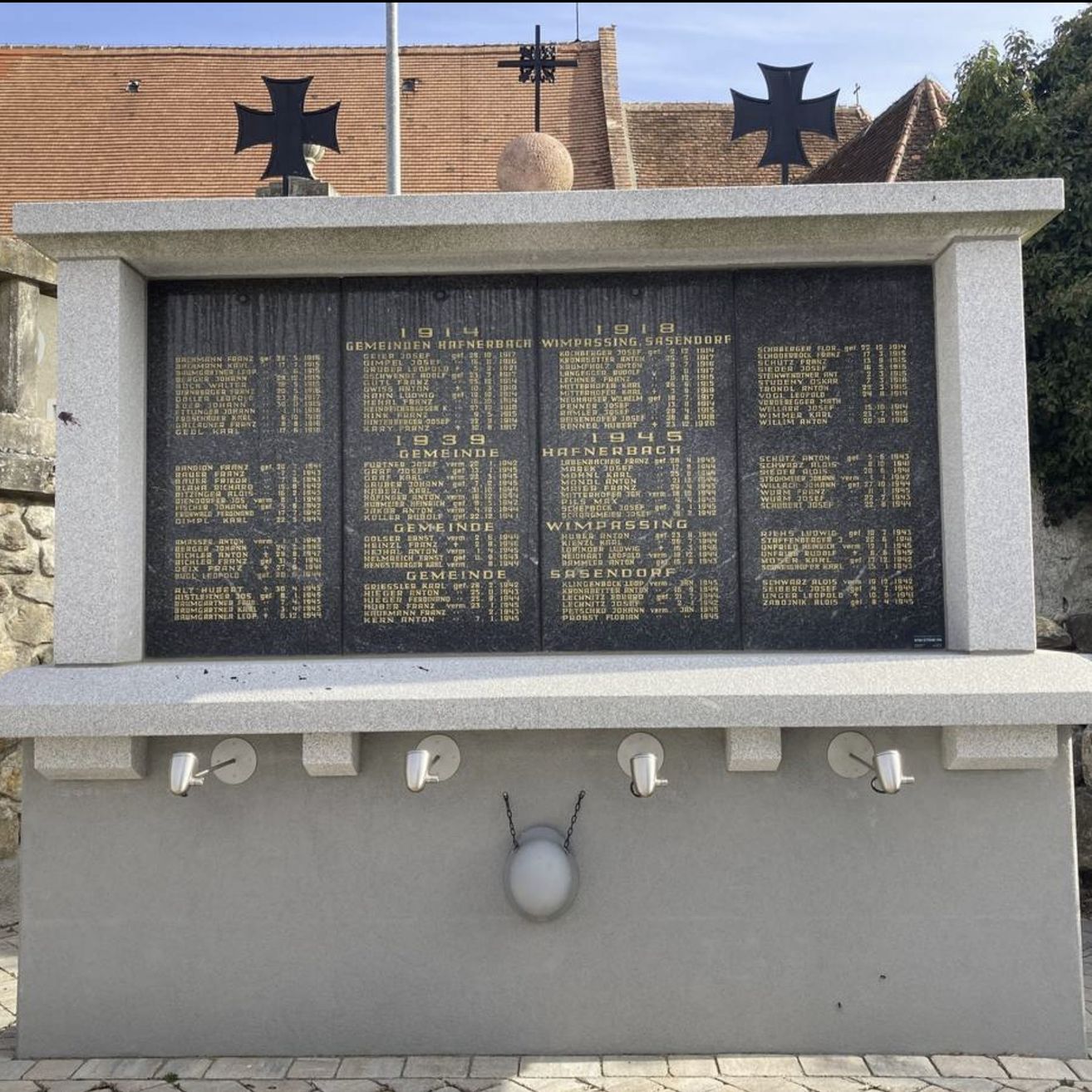 Ein Denkmal zeigt Namen und Daten in Deutsch, flankiert von zwei schwarzen Kreuzen. Darunter sind drei silberne Lampen befestigt. Hinter dem Denkmal steht ein Gebäude mit einem roten Dach und Grünpflanzen.