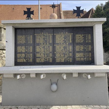Ein Denkmal zeigt Namen und Daten in Deutsch, flankiert von zwei schwarzen Kreuzen. Darunter sind drei silberne Lampen befestigt. Hinter dem Denkmal steht ein Gebäude mit einem roten Dach und Grünpflanzen.
