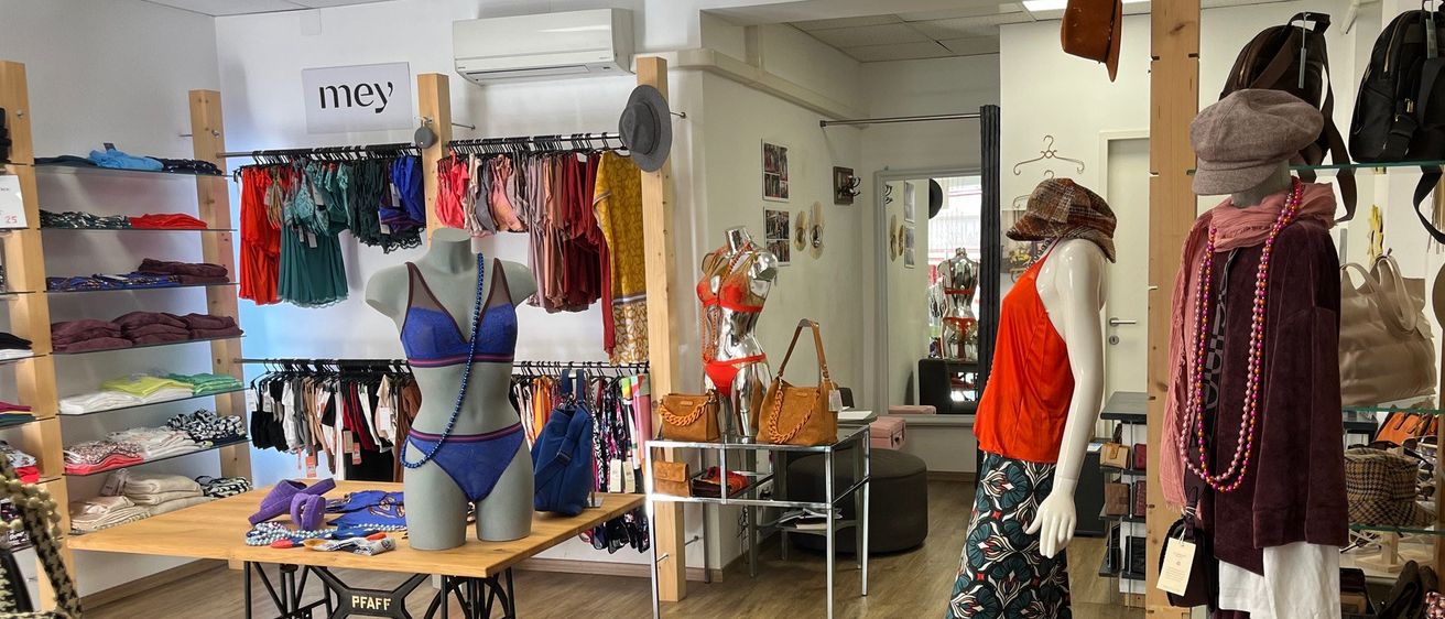 Ein Boutique zeigt eine Vielzahl von Kleidungsstücken und Accessoires an Mannequins und Regalen. Die Mannequins sind in bunten Outfits, Hüten und Schals gekleidet. Der Boden ist aus Holz und auf einem Tisch steht eine Nähmaschine.