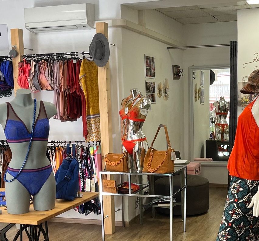 Ein Boutique zeigt eine Vielzahl von Kleidungsstücken und Accessoires an Mannequins und Regalen. Die Mannequins sind in bunten Outfits, Hüten und Schals gekleidet. Der Boden ist aus Holz und auf einem Tisch steht eine Nähmaschine.