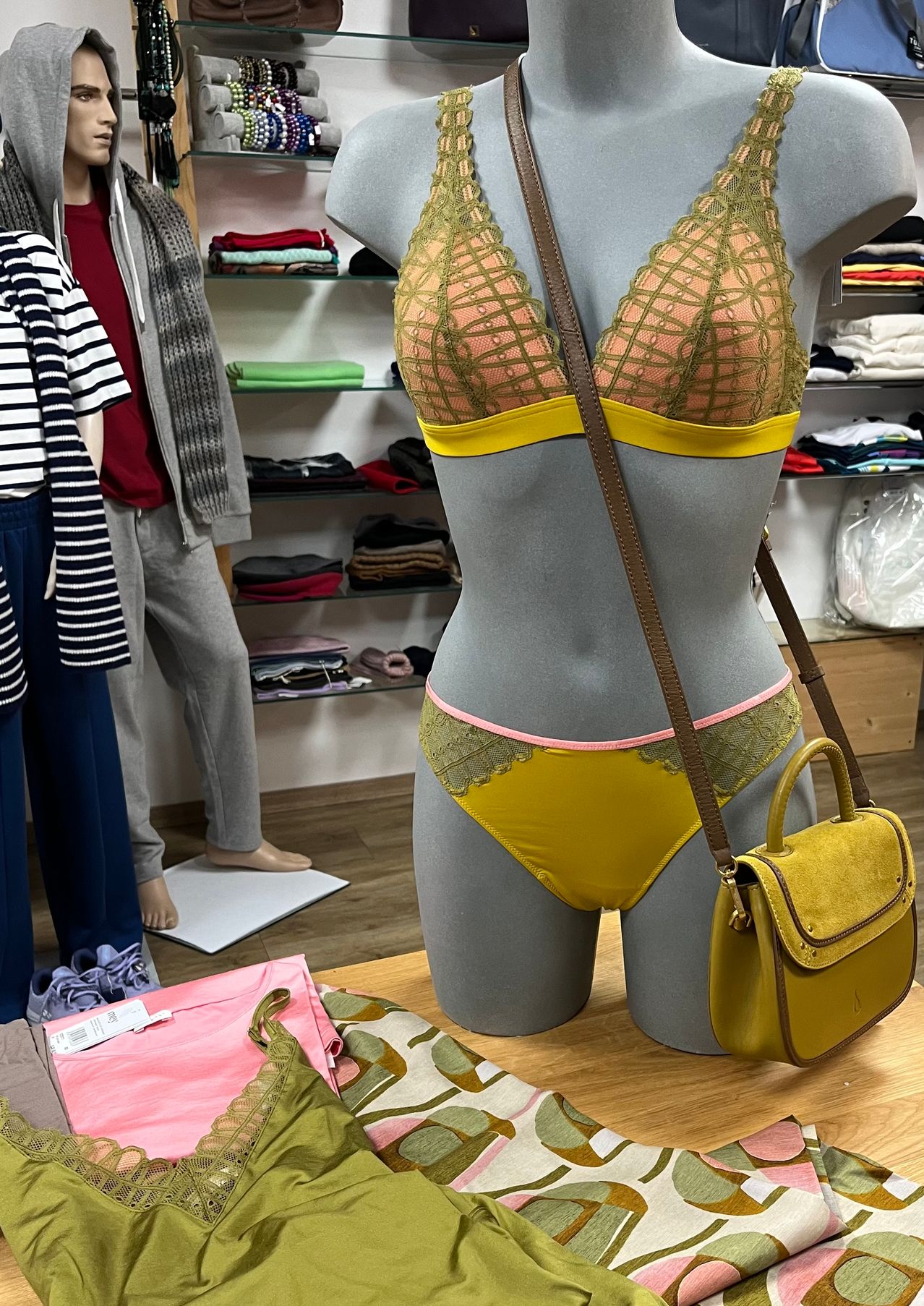 Eine Puppe mit gelber Unterwäsche und einer braunen Umhängetasche steht in einem Bekleidungsgeschäft. Dahinter sind Kleider an den Regalen. Eine Person in Rot und Grau ist in der Nähe.