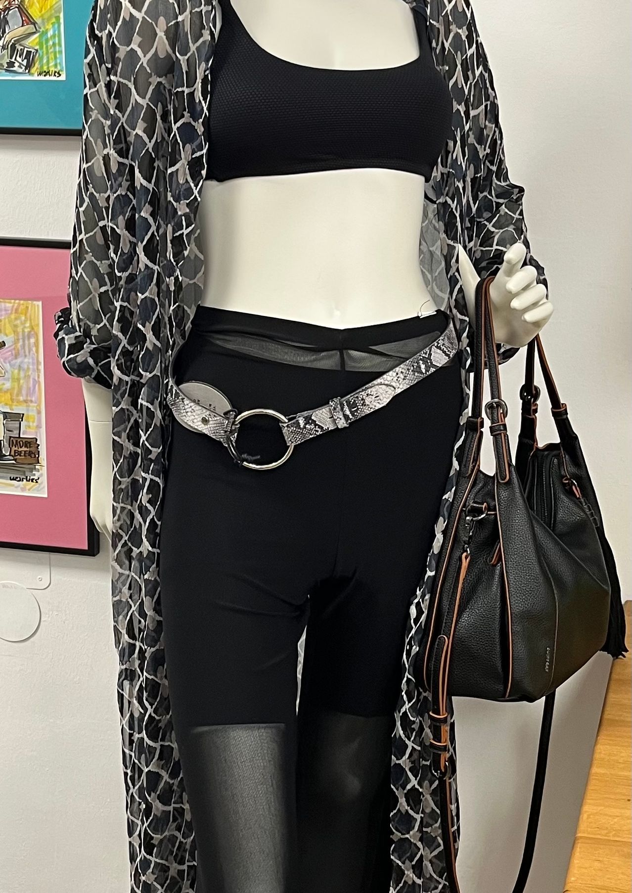 Eine Puppe in einem schwarzen Oberteil und Hose mit einem großen Schlangenmustergürtel, der eine schwarze Handtasche hält. Der Hintergrund zeigt einen rosa Rahmen mit abstrakter Kunst.