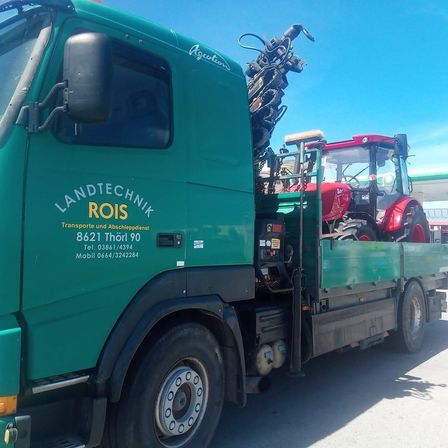 Ein grüner LKW mit 'LANDTECHNIK ROIS' auf der Seite ist auf einem Parkplatz geparkt. Er transportiert einen roten Traktor auf seinem Ladeflächen.