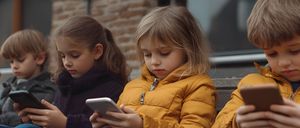 Ein kleines Mädchen in einer gelben Daunenjacke schaut auf ihr Smartphone, während es neben einem anderen Mädchen an einer Ziegelwand sitzt.