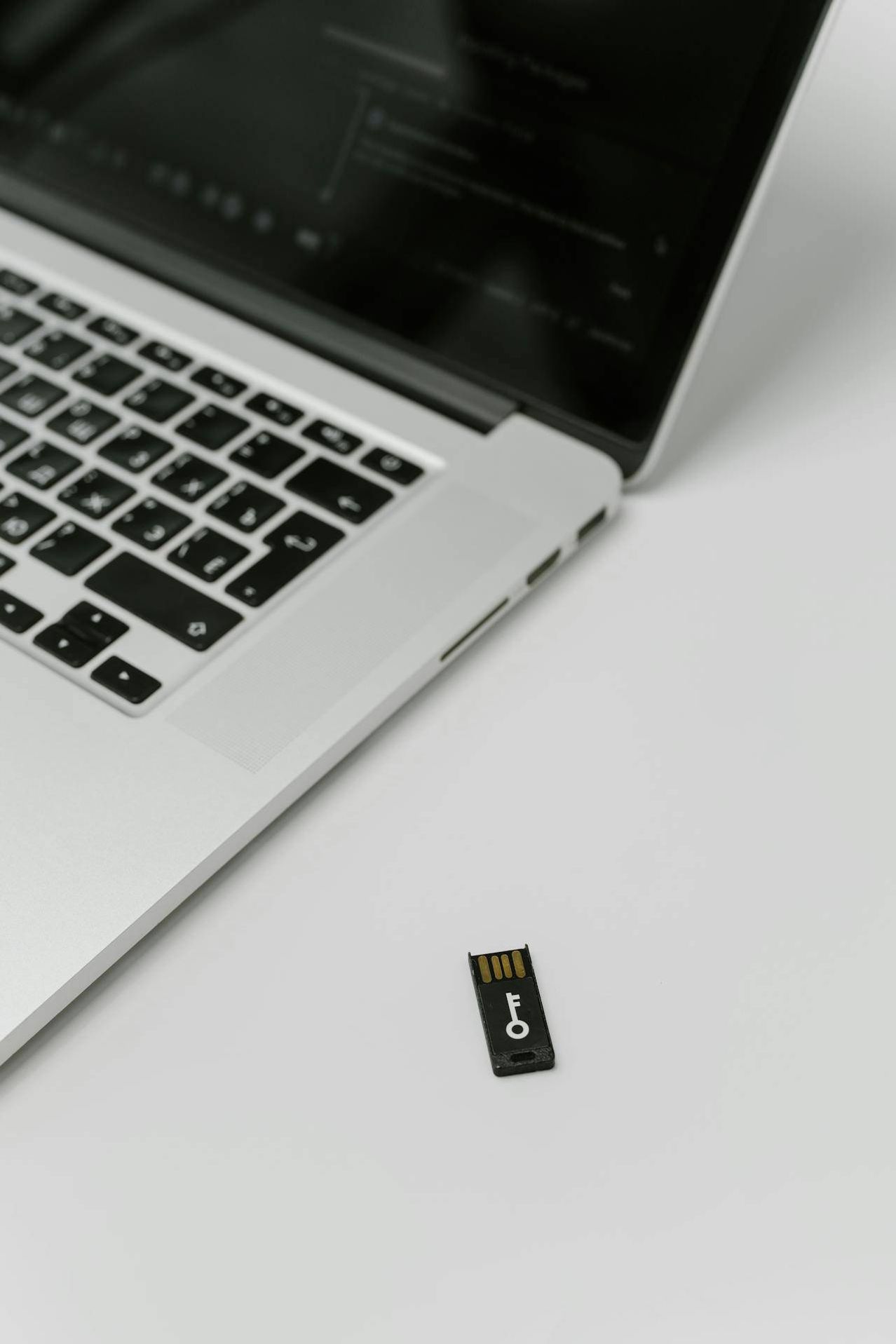 Ein silberner Laptop mit schwarzer Tastatur liegt auf einer weißen Oberfläche, mit einem kleinen USB-Stick daneben.