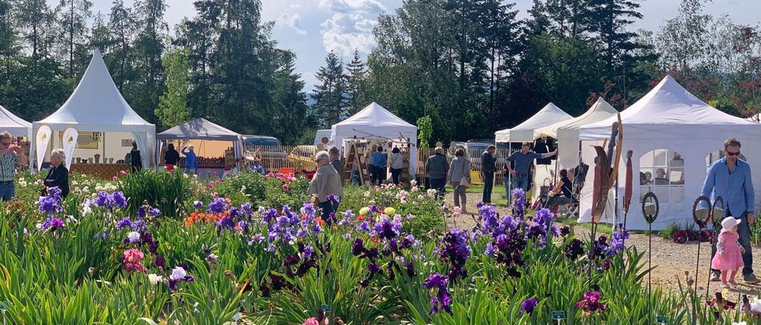 Ein Gartenmarkt mit Besuchern, umgeben von weißen Zelten und farbenfrohen Blumen, unter einem bewölkten Himmel mit hohen Bäumen im Hintergrund.