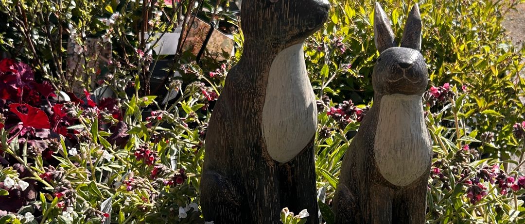 Zwei hölzerne Kaninchenstatuen stehen in einem Garten, umgeben von verschiedenen Blumen und Pflanzen. Ein Osterei ist ebenfalls in der Nähe vorhanden.