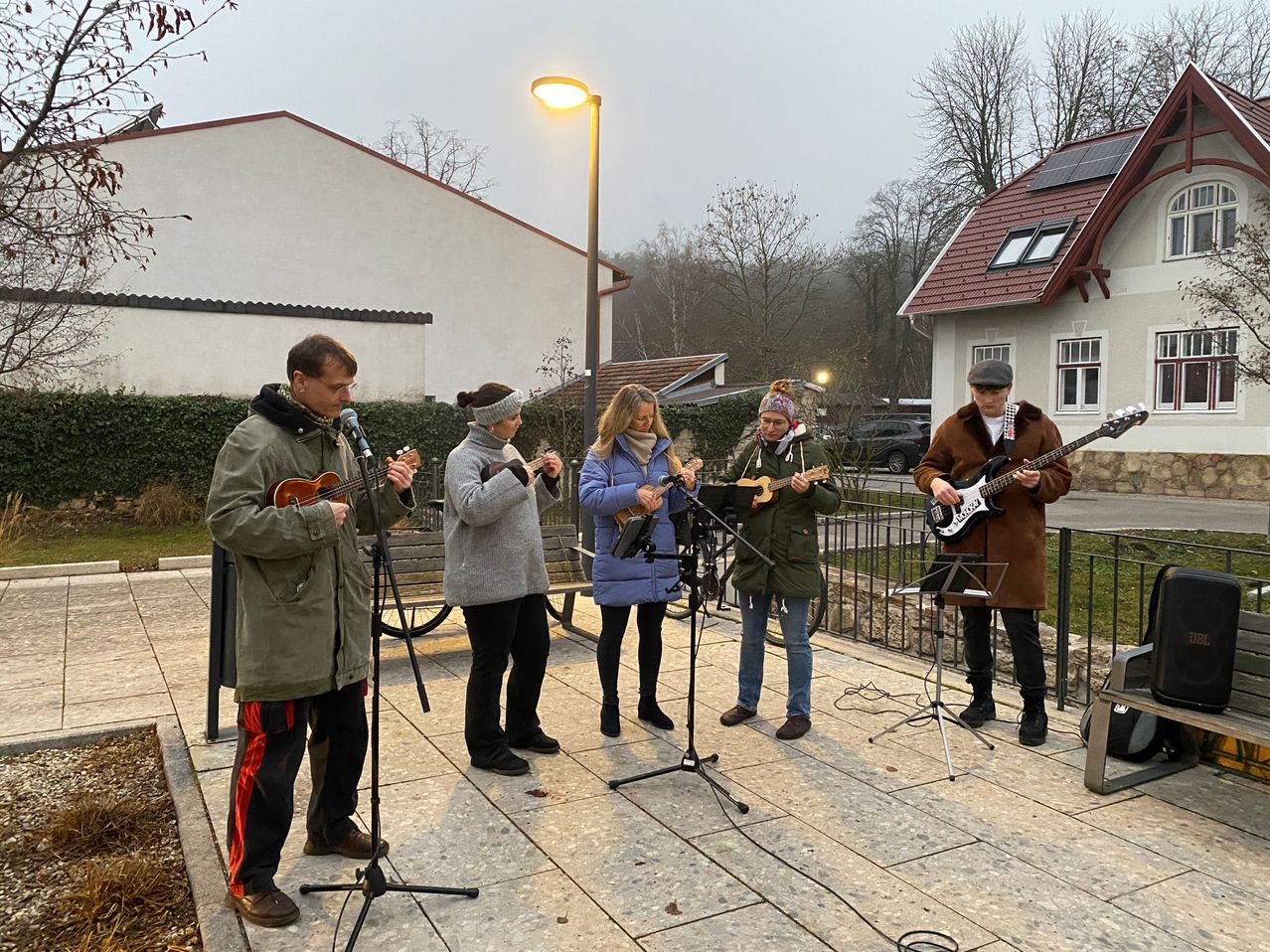 Fünf Musiker spielen auf einer gefliesten Bühne in einem Wohngebiet. Sie spielen Gitarren und Ukulelen. Hinter ihnen ist eine Straßenlaterne und ein Gebäude mit Solarmodulen zu sehen.