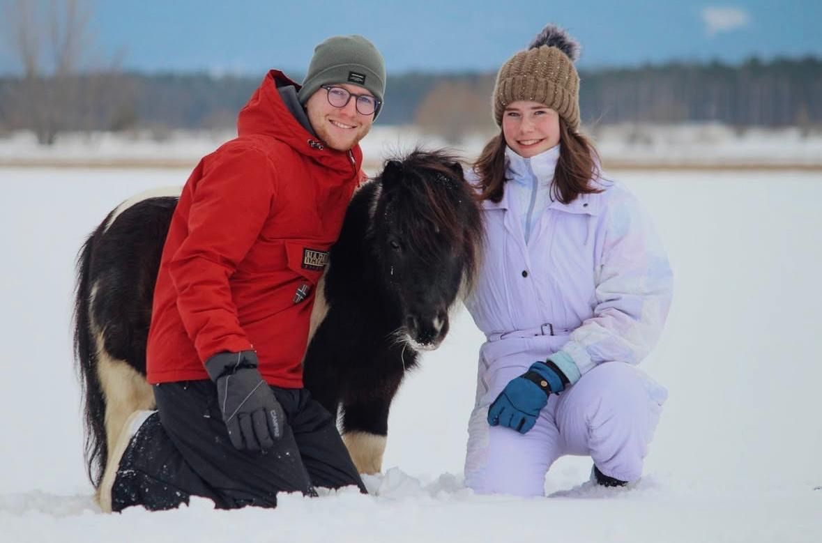 Ein Paar in Winterkleidung posiert mit einem kleinen Pferd in einem verschneiten Feld, beide lächeln.