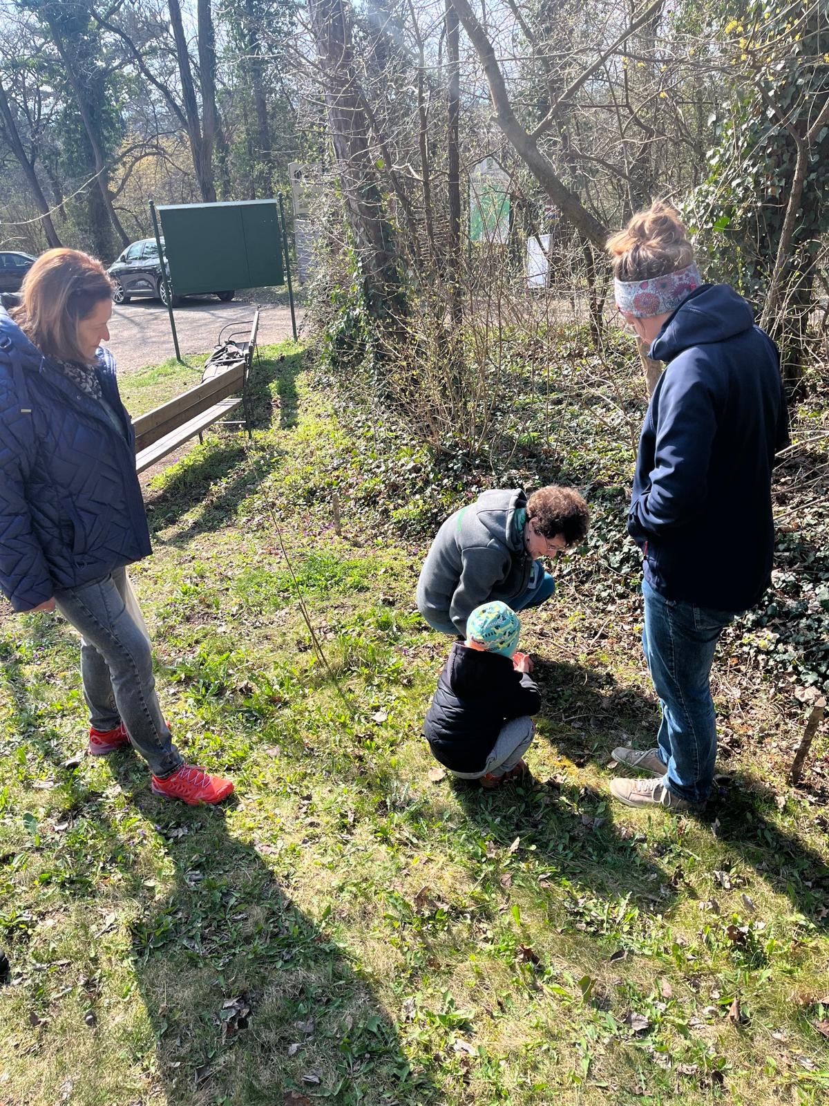 Eine Gruppe von vier Menschen ist draußen, in der Nähe eines Baumes. Ein Kind kniet auf dem Boden, während ein Mann und eine Frau zusehen.