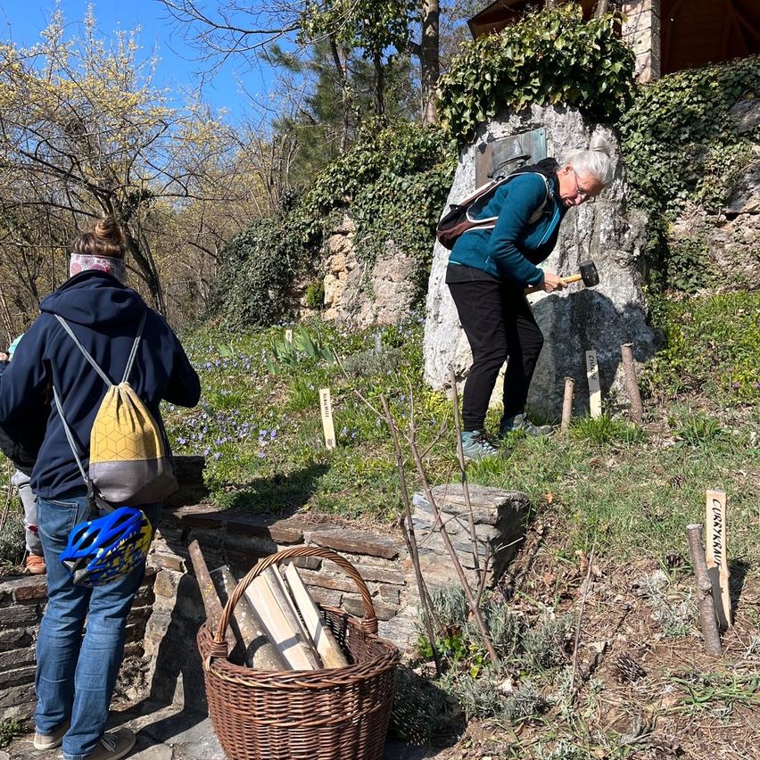Zwei Personen arbeiten in einem Garten, eine mit einem Hammer, die andere mit einem Rucksack. Ein Korb mit Holz ist in der Nähe.