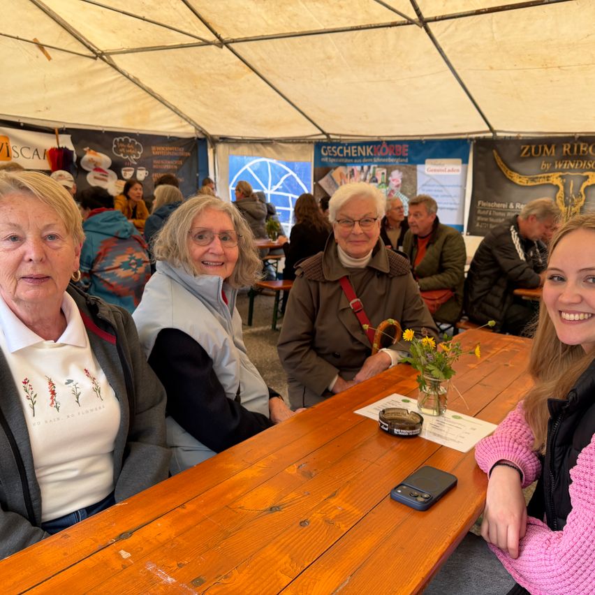 Four people are seated at a table under a tent. Two older women wear glasses and jackets. A man and a young woman sit across from them. A table holds a cell phone, flowers, and a document.