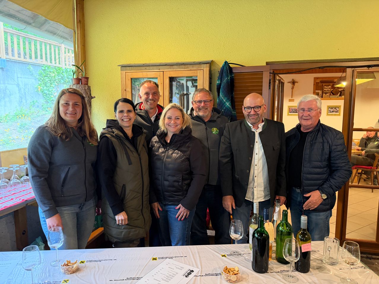 A group of smiling adults stands in a room with a table full of wine bottles and glasses. A woman is on the left, and a man is on the right. The room has yellow walls and a window.