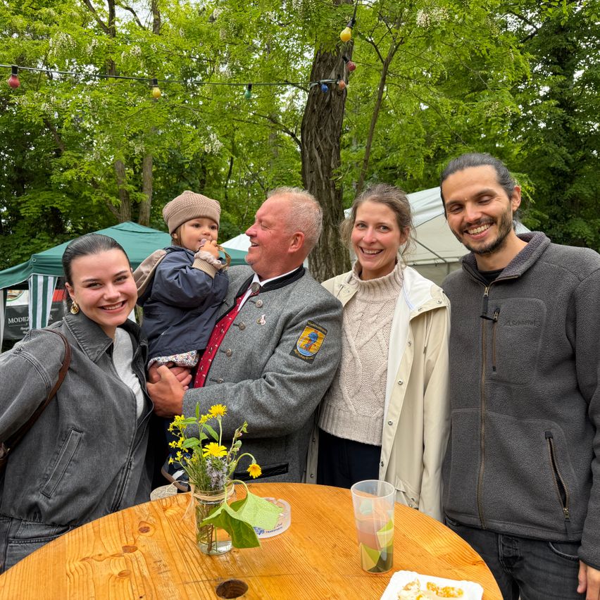 Five people are smiling for a photo in an outdoor setting. A child is being held by an older man, and a woman holds flowers. A wooden table and a cup with a drink are in the foreground.