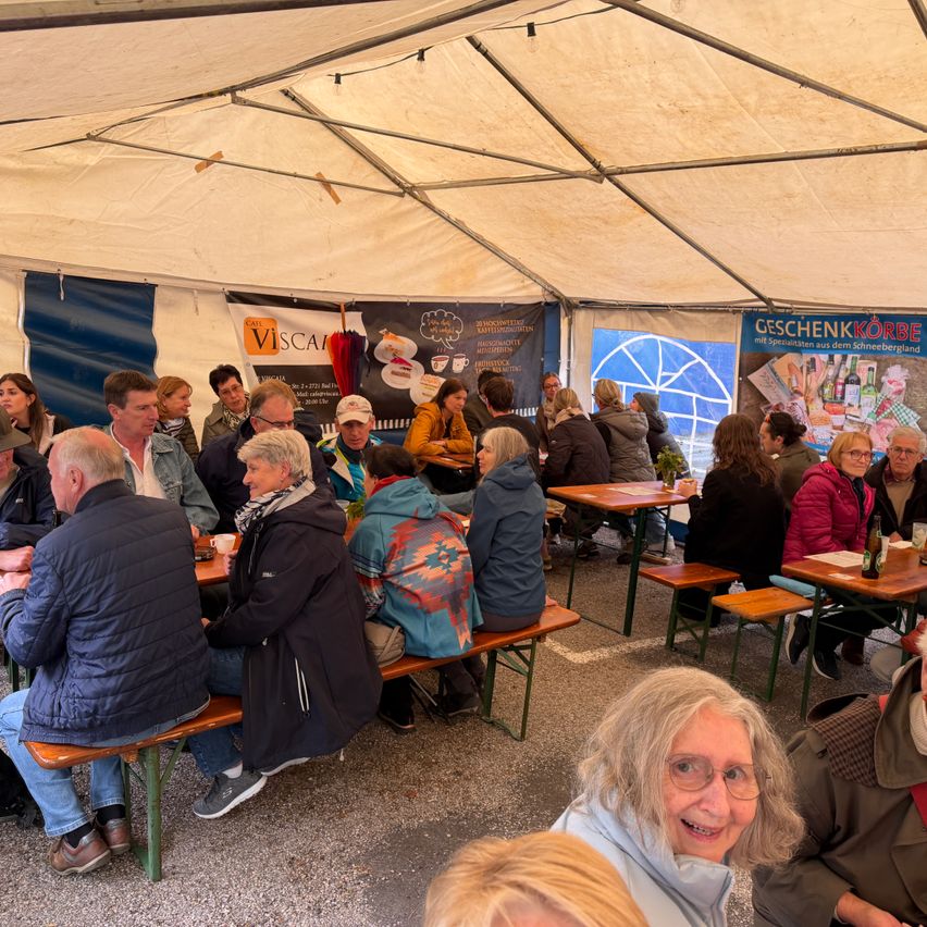 A group of adults is seated around tables in a tent. Some tables have bottles, cups, and plates. There are banners with text in the background.