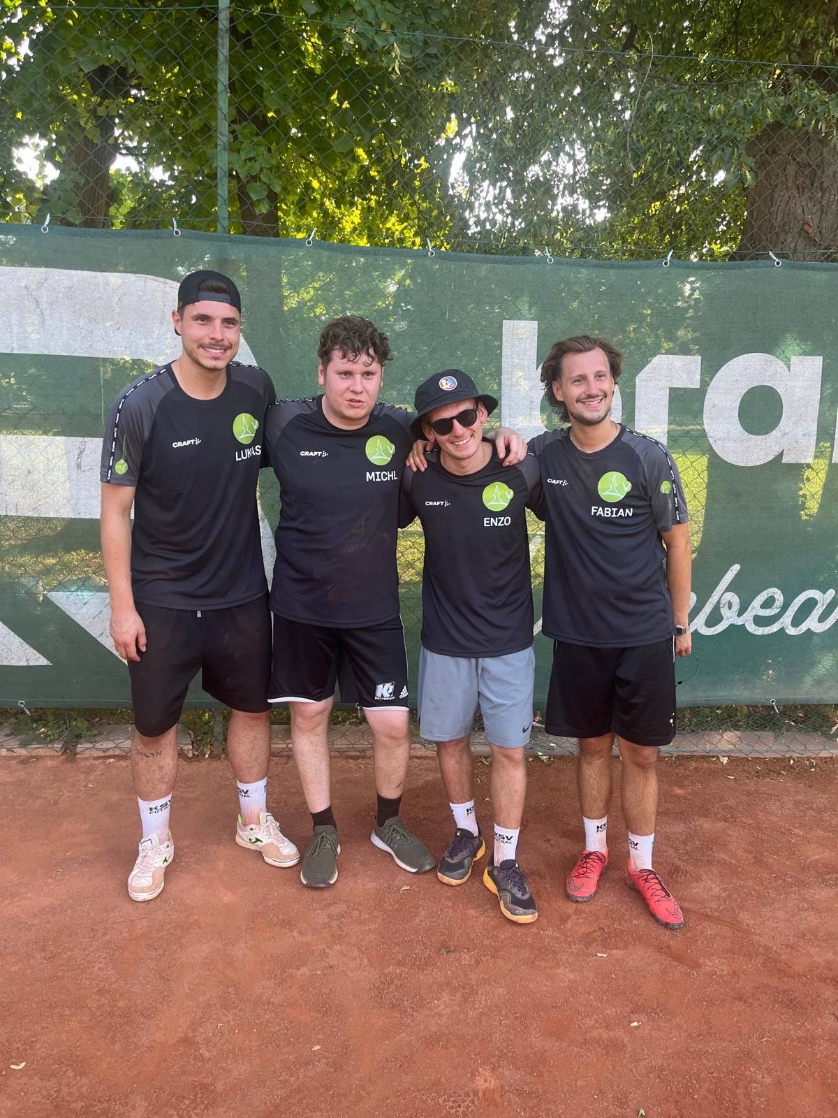 Fünf Männer stehen auf einem Tennisplatz und tragen passende schwarze Trikots. Zwei von ihnen haben Sonnenbrillen auf. Hinter ihnen befindet sich ein grüner Banner mit weißen Buchstaben.