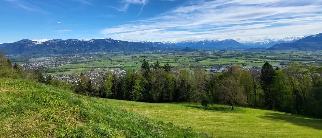 Ein landschaftlicher Blick von einem Hügel mit einem üppigen grünen Tal darunter, Bergen in der Ferne und einem klaren blauen Himmel mit Wolken.
