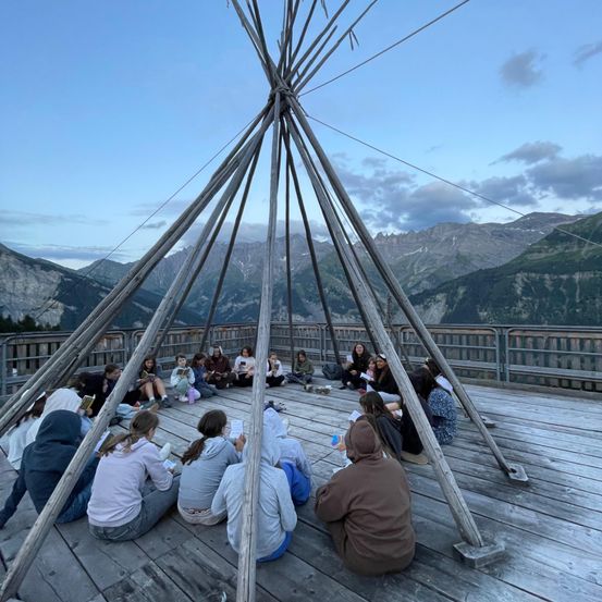 Eine Gruppe von Menschen sitzt im Kreis unter einer großen Holzstruktur, möglicherweise einem Tipi, auf einer Holzplattform mit Blick auf Berge im Hintergrund.