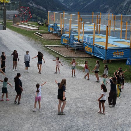 Eine Gruppe von Kindern und Erwachsenen versammelt sich in einem Außenbereich, möglicherweise einem Spielplatz, mit einem blauen Trampolin und einer roten Struktur im Hintergrund.