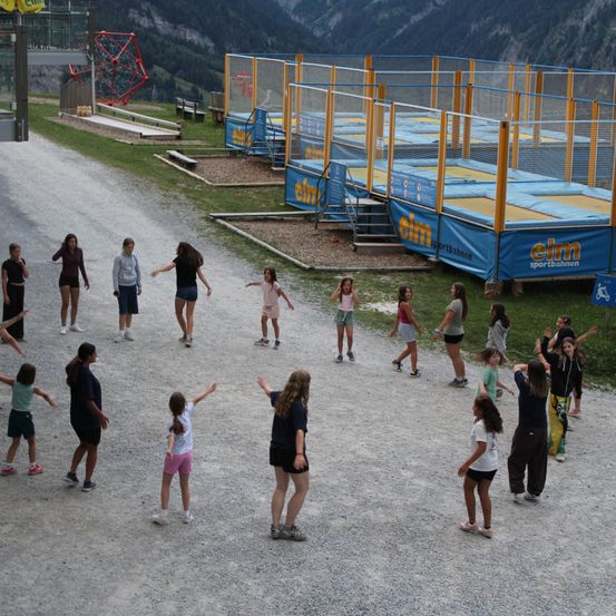 Eine Gruppe von Kindern und Erwachsenen versammelt sich in einem Außenbereich, möglicherweise einem Spielplatz, mit einem blauen Trampolin und einer roten Struktur im Hintergrund.
