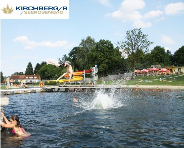 Eine Person taucht in einen See bei Kirchberg/Seerosenbad ein, umgeben von Bäumen und Menschen, die den Tag genießen.