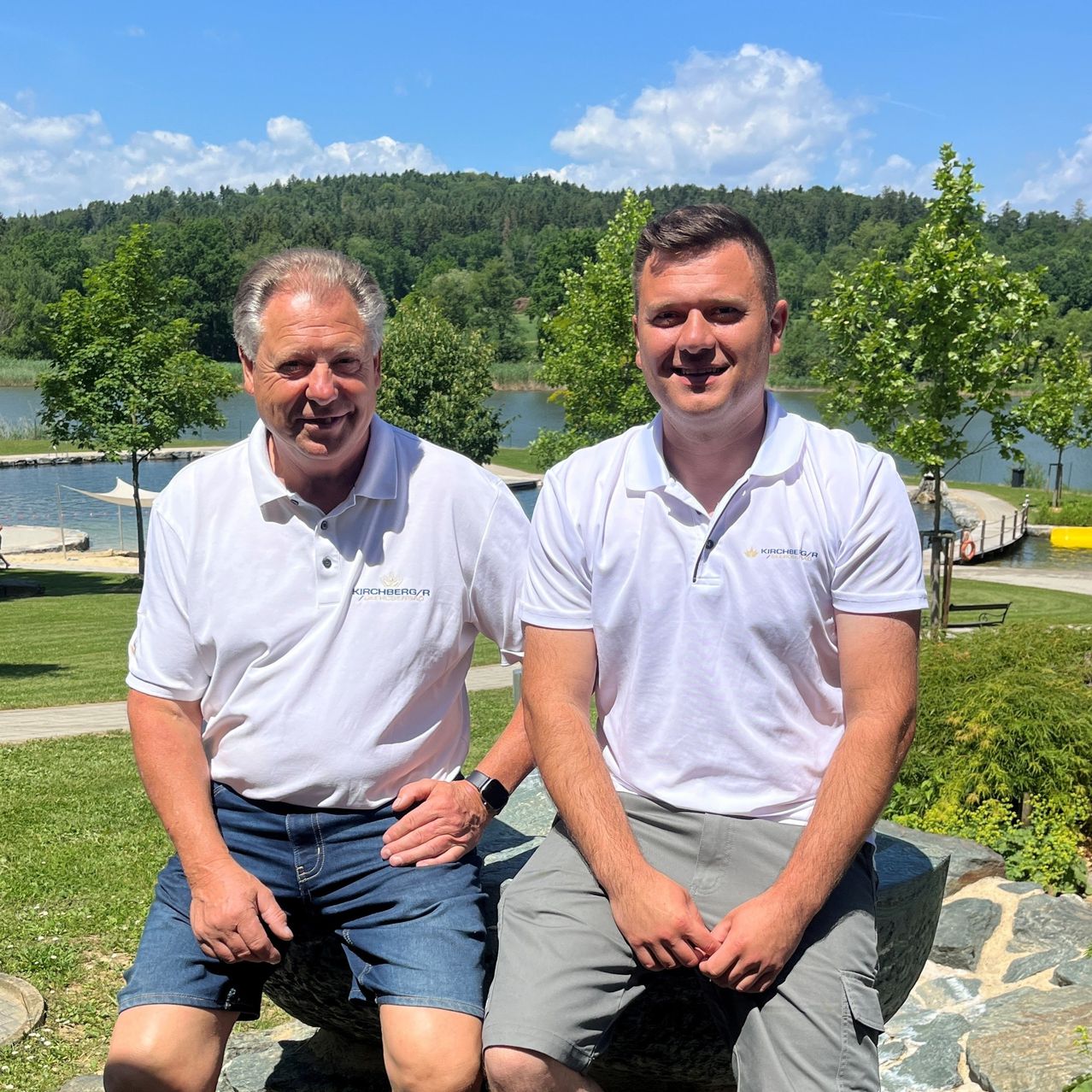 Two men, both in white polo shirts, sit on a rock in a park by the lake, smiling. Trees and grass surround them, and the sky is clear.