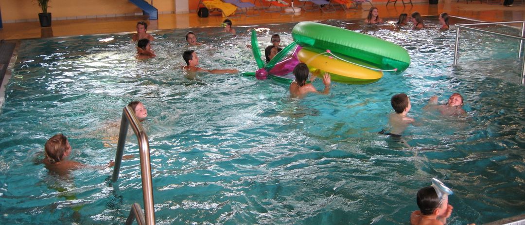 Mehrere Kinder schwimmen in einem Innenpool, einige benutzen aufblasbare Spielzeuge. Im Hintergrund sind farbenfrohe Liegestühle und ein Regenschirm zu sehen.