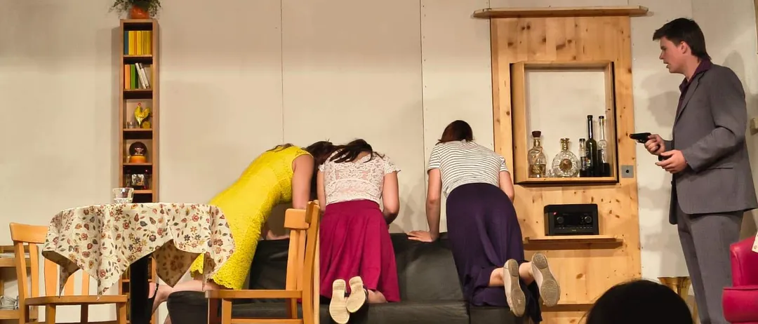 Drei Frauen beugen sich über eine Couch in einer inszenierten Szene, während eine Person im Hintergrund eine Waffe hält.
