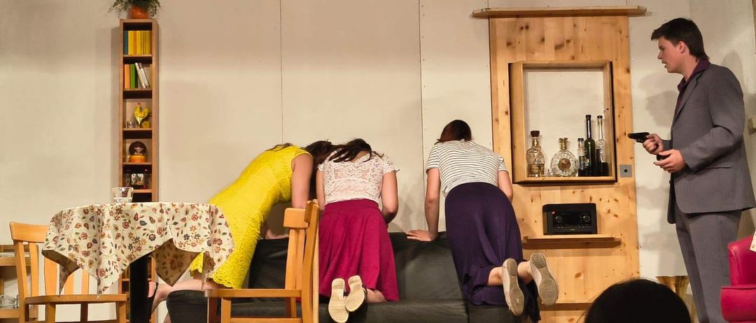 Drei Frauen beugen sich über eine Couch in einer inszenierten Szene, während eine Person im Hintergrund eine Waffe hält.