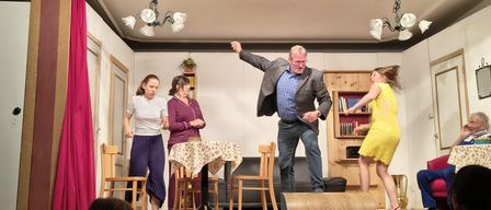 A group of people are on a stage performing a skit. A man in a suit is gesturing while a woman in a yellow dress is running. There are chairs, a table, and a couch in the background.