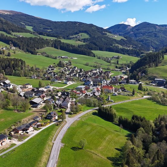 Luftaufnahme eines malerischen Dorfes, umgeben von saftig grünen Feldern und dichten Wäldern, eingebettet in den Bergen unter einem blauen Himmel mit Wolken.