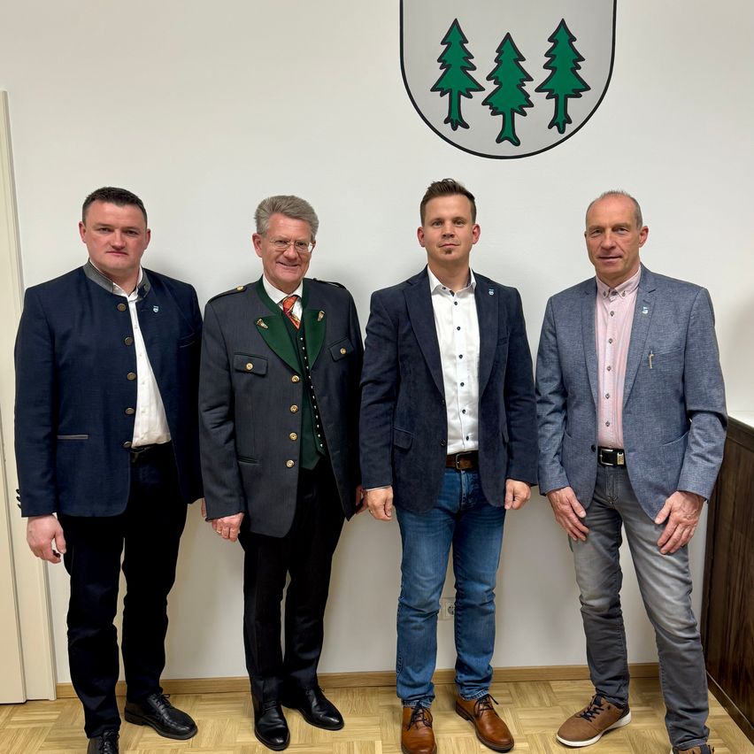 Vier Männer stehen zusammen vor einer weißen Wand mit einem Logo, das drei grüne Bäume zeigt. Sie sind in verschiedenen Arten von Anzügen gekleidet, ein Mann trägt eine traditionelle Uniform.