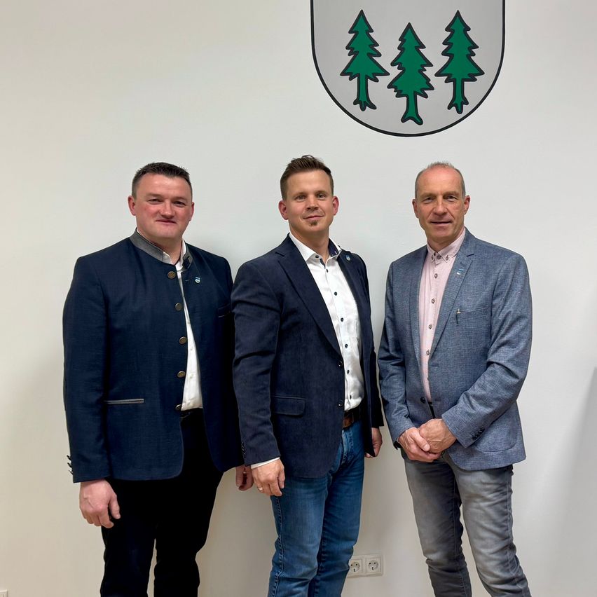 Drei Männer stehen zusammen vor einer weißen Wand. Sie sind in formeller Kleidung gekleidet. Der Mann links trägt einen dunklen Anzug, der Mann in der Mitte einen helleren Blazer und der Mann rechts ein rosa Hemd. Über ihnen befindet sich ein weißes Emblem mit drei grünen Bäumen.