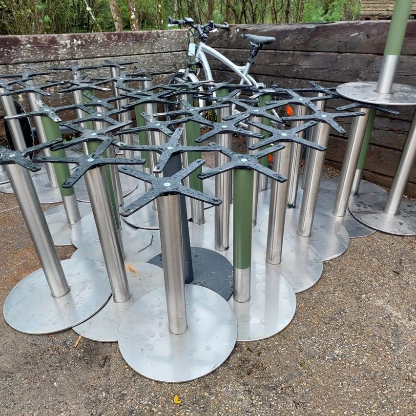 Mehrere Edelstahl-Tischbeine sind in einer Außenumgebung auf dem Boden angeordnet. Dahinter ist ein Fahrrad an einer Holzwand geparkt.