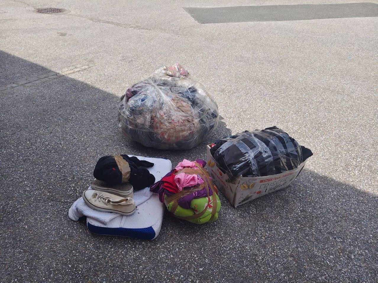 Verschiedene Taschen und Kisten mit Kleidung und Schuhen sind auf dem Boden platziert. Einige Gegenstände sind in Plastik eingewickelt.