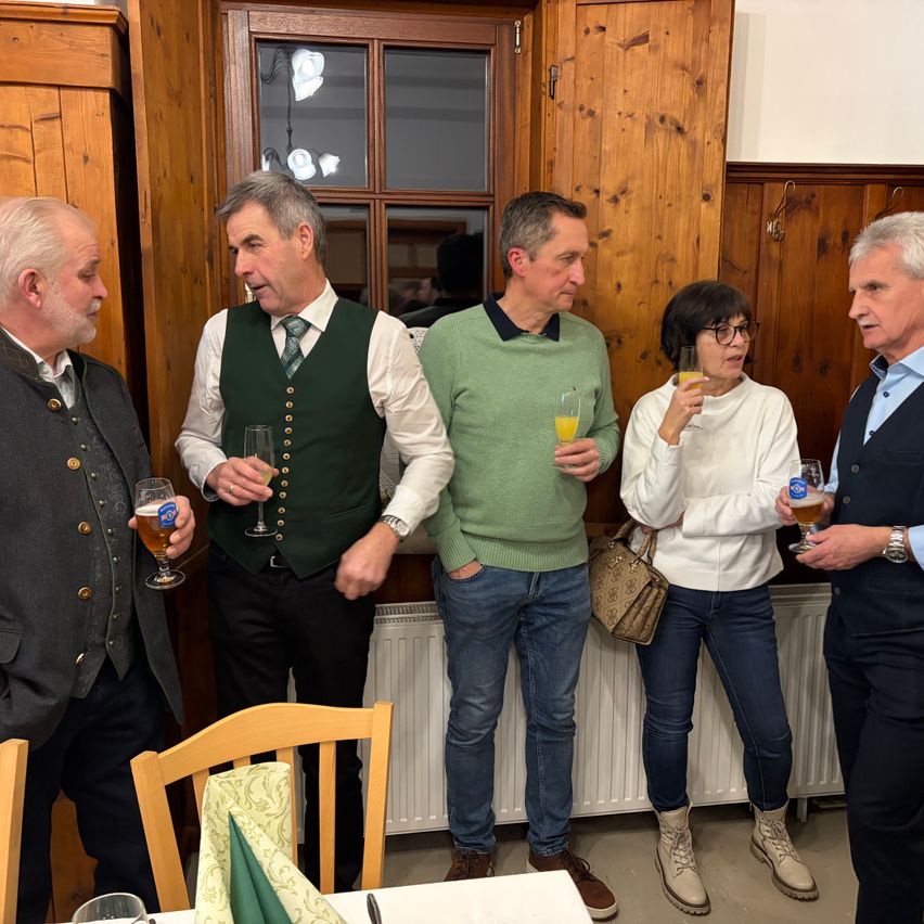 Fünf Erwachsene stehen in einem Raum mit Holzwänden. Drei Männer und zwei Frauen halten Gläser mit Bier. Die Männer tragen grüne Westen, ein grünes Sweatshirt und eine dunkle Weste. Die Frauen tragen ein weißes Sweatshirt und eine Brille.