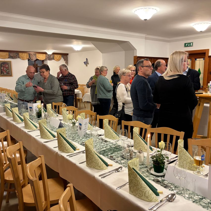 Eine Gruppe von Menschen steht um einen langen Esstisch in einem Restaurant. Der Tisch ist mit Blumen, Kerzen und Weingläsern gedeckt. Einige Leute schauen auf ihre Telefone.