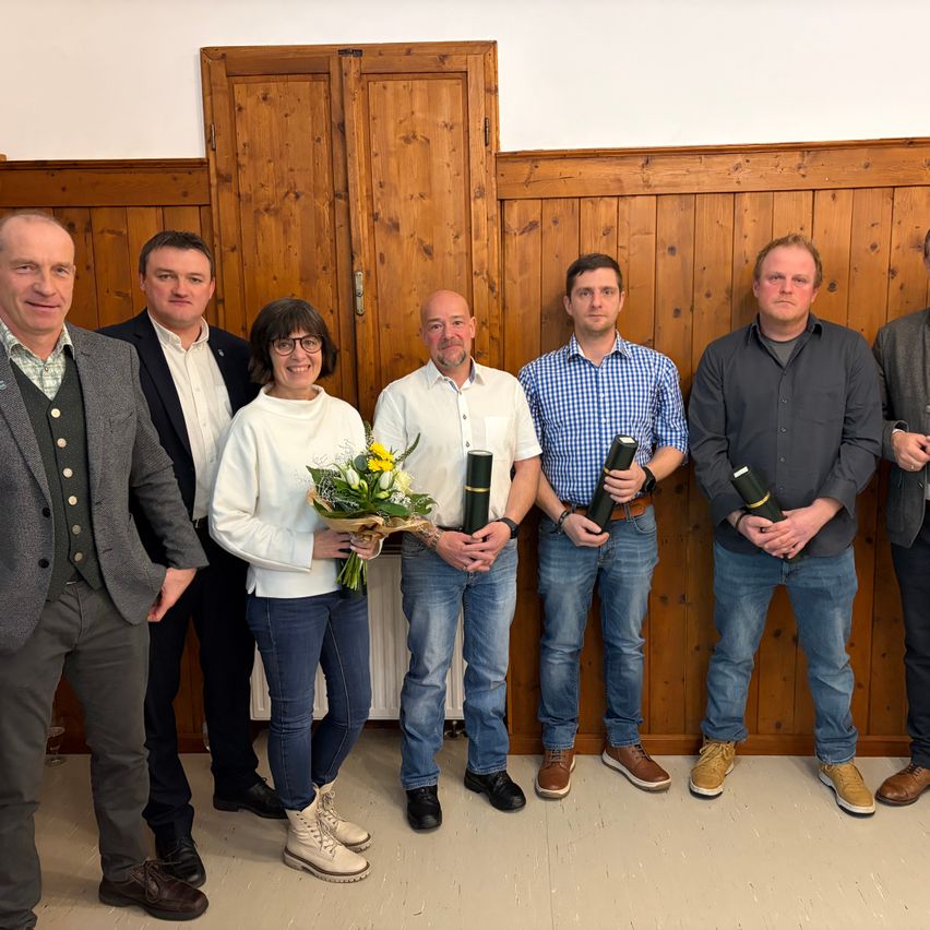 Eine Gruppe von Menschen steht in einem Raum und posiert für ein Foto. Eine Frau in der Mitte hält einen Blumenstrauß. Der Mann neben ihr hält ein schwarzes zylindrisches Objekt. Sie alle scheinen für ein Gruppenfoto zu posieren.