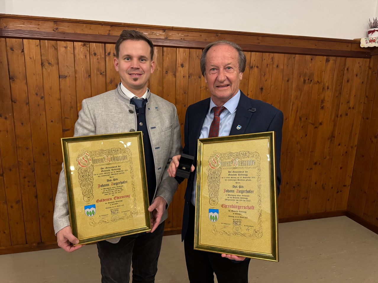 Zwei Männer stehen vor einer Holzwand, jeder hält ein gerahmtes Zertifikat in den Händen. Der Mann auf der linken Seite lächelt, trägt einen hellgrauen Anzug und eine Krawatte. Der Mann auf der rechten Seite trägt einen dunklen Anzug und eine rote Krawatte und hält sein Zertifikat stolz in die Höhe.