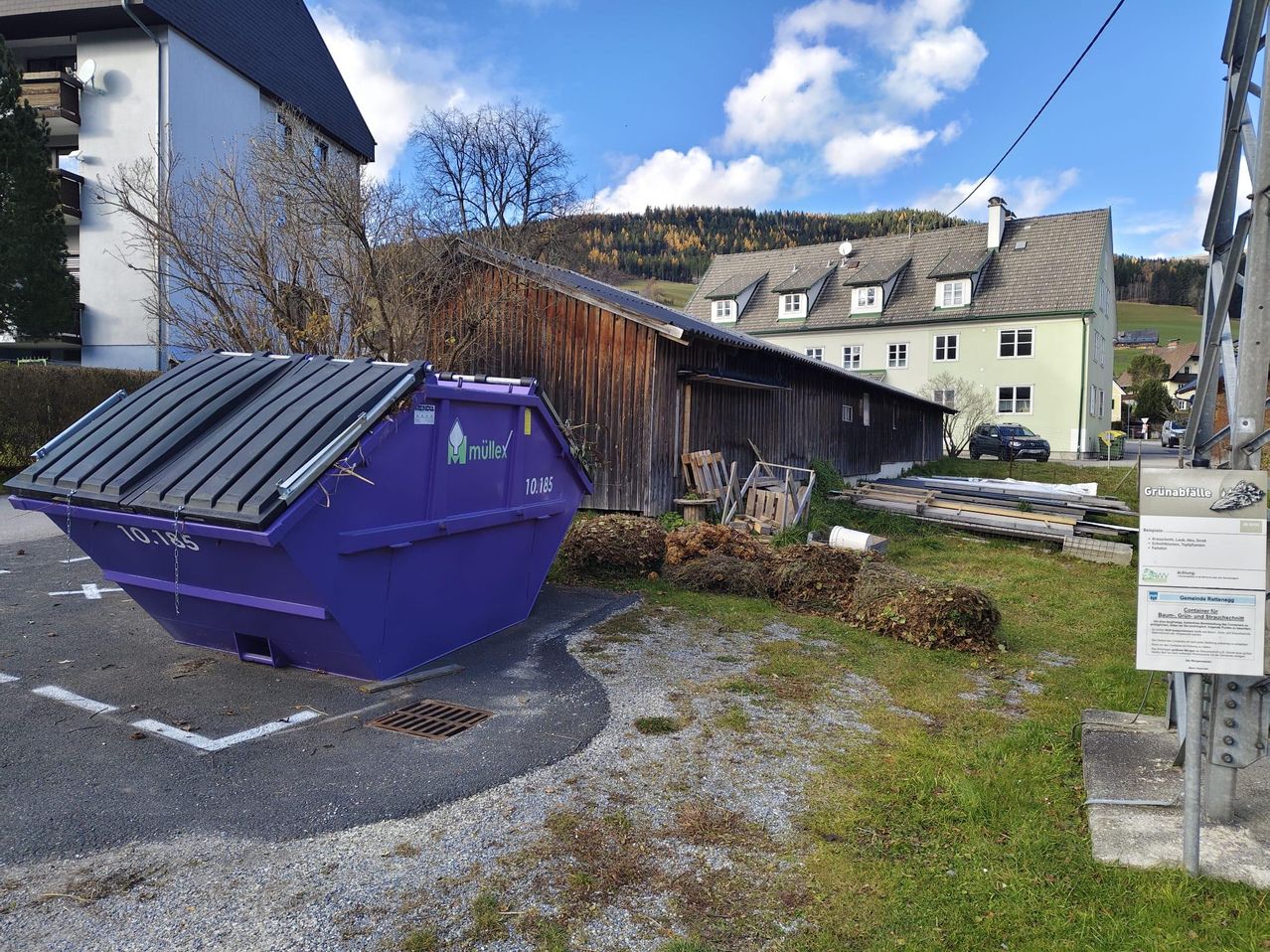 Ein lila Müllcontainer steht vor einem Haus, ein Auto ist in der Nähe geparkt und ein Schuppen steht hinter dem Haus. Das Gebiet ist von Bäumen umgeben und der Himmel ist blau mit Wolken.